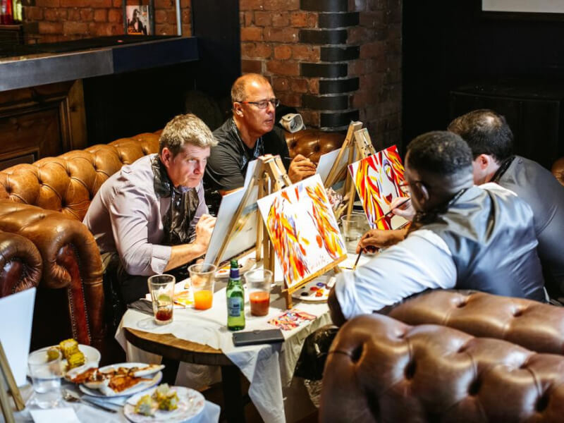 Four people sitting in leather booth chairs, painting on canvas